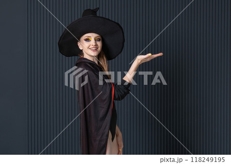Attractive smiling woman in witch hat shows something with her palms against dark wall. Halloween party. Black friday. Space for text Attractive smiling woman in witch hat shows something with her palms against dark wall. Halloween party. Black friday. Space for text 118249195