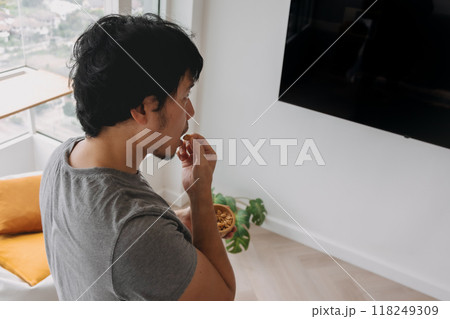 Man standing and eating peanuts snack while watching television in the room. 118249309