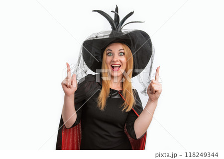 Portrait of young woman with green eyes and black witch hat pointing her fingers up. Halloween party, white background. Young witch 118249344