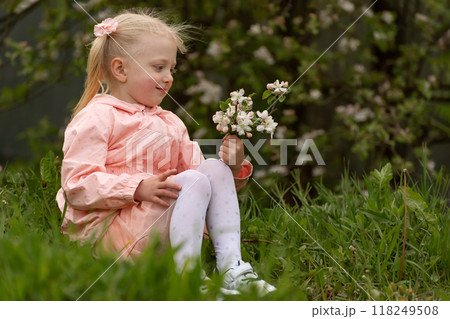 Blonde girl with two tails sits outside in the spring. Child in the blooming garden with flowers in her hands. 118249508