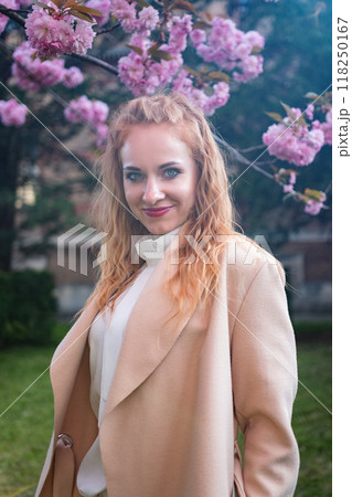 Portrait of attractive young woman in stylish beige coat near blossoming pink tree in park in early spring. Fashion model with red wavy hair. Portrait of attractive young woman in stylish beige coat near blossoming pink tree in park in early spring. Fashion model with red wavy hair. 118250167