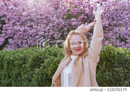 Young beautiful woman smiling and standing in blooming sakura park raised her hands. Happy gentle woman in blossoming park. 118250174