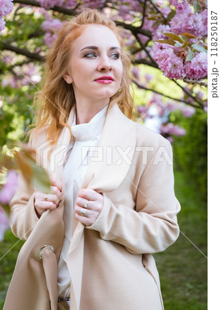 Portrait of pretty young woman dressed stylish beige coat posing near blossoming pink tree in park in early spring. Fashion model with red wavy hair. Portrait of pretty young woman dressed stylish beige coat posing near blossoming pink tree in park in early spring. Fashion model with red wavy hair. 118250187