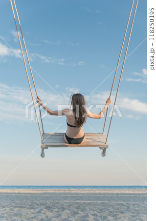 Young slim girl in swimsuit pushes on rope swing on blue sky and beach background. Vertical frame. Young slim girl in swimsuit pushes on rope swing on blue sky and beach background. Vertical frame. 118250195