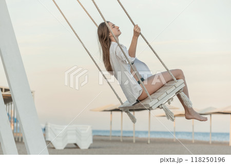 Happy beautiful young girl in white clothes pushes in swing on sky background. Beautiful soft sunset light. Pretty girl on the beach Happy beautiful young girl in white clothes pushes in swing on sky background. Beautiful soft sunset light. Pretty girl on the beach 118250196