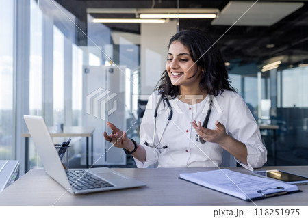 Young female doctor uses laptop for video call, female doctor talks consults patients remotely. He happily smiles, works remotely inside the clinic office, communicates and helps people. 118251975