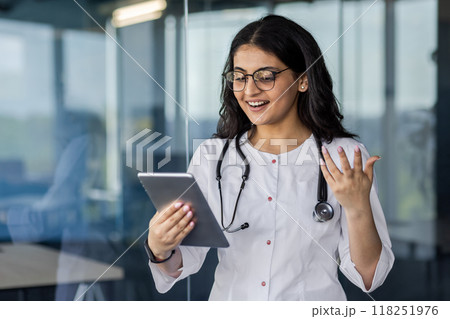 Indian woman doctor doctor inside clinic using tablet computer for video call, online communication consulting with patient remotely, female worker in white medical coat with stethoscope. 118251976