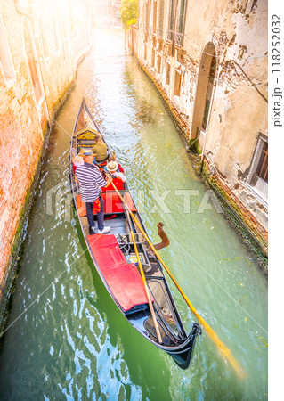 A gondola glides through a peaceful canal in Venice, Italy, as the sun casts a warm glow. The gondolier navigates while passengers enjoy the picturesque surroundings of historic buildings. 118252032