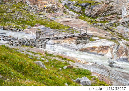 A wooden bridge spans a rushing stream sourced from the Schlaten Glacier, surrounded by rocky terrain and vibrant green vegetation in Hohe Tauern National Park. 118252037