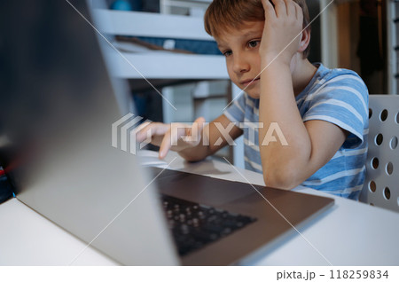 caucasian little boy wearing wireless headphones studying at home typing on laptop 118259834