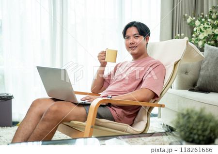 An Asian man is enjoying coffee while working on his laptop from a lounge chair in the living room. An Asian man is enjoying coffee while working on his laptop from a lounge chair in the living room. 118261686