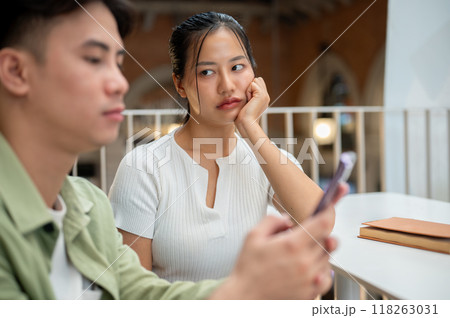An unhappy Asian woman is feeling jealous, looking at her boyfriend while he is texting on his phone An unhappy Asian woman is feeling jealous, looking at her boyfriend while he is texting on his phone 118263031