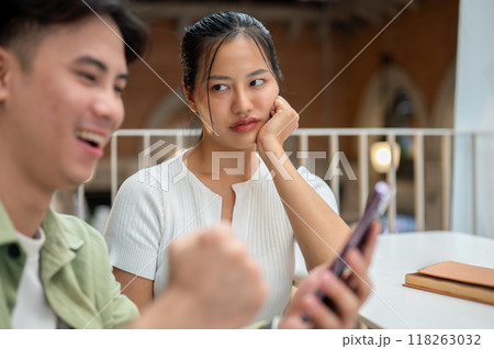 An unhappy Asian woman is feeling jealous, looking at her boyfriend while he is texting on his phone 118263032
