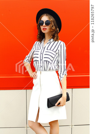 Beautiful young woman model wearing white striped shirt, black handbag clutch, round hat, skirt in the city Beautiful young woman model wearing white striped shirt, black handbag clutch, round hat, skirt in the city 118265797
