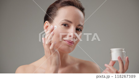 Beauty, health, cosmetics, anti-aging therapy and skin care concept - young beautiful brunette Caucasian woman holds a jar of moisturizer in her hands and applies the cream to her face skin 118267389