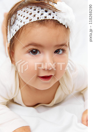 Tiny tot in cute outfit peeks over blanket on soft bed with white bedding, smiling happily, her face full of joy against studio background. Tiny tot in cute outfit peeks over blanket on soft bed with white bedding, smiling happily, her face full of joy against studio background. 118268290
