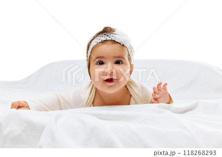 Little baby in white onesie and headband looks curiously at camera, crawling on soft white bed with focused expression. Little baby in white onesie and headband looks curiously at camera, crawling on soft white bed with focused expression. 118268293