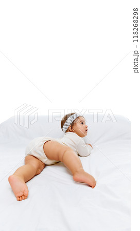 Infant in white outfit crawls on white bed, glancing over her shoulder with wide, curious eyes, focused on something in distance. 118268298
