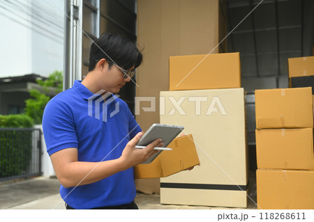 Delivery man holding a cardboard box and checking or input information on digital tablet next to a vehicle loaded with package 118268611
