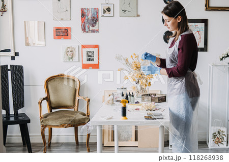 Young woman preparing resin art in a creative studio, surrounded by dried flowers and artistic decor. Home crafting, DIY projects, resin art, handmade decor, creative workspace. 118268938