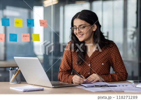 Happy woman financier doing paperwork inside office, latin american smiling pleased preparing financial report, accountant recording data and filling forms sitting at table with documents and laptop. Happy woman financier doing paperwork inside office, latin american smiling pleased preparing financial report, accountant recording data and filling forms sitting at table with documents and laptop. 118271289