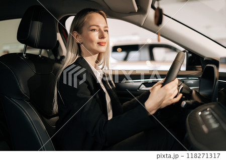 Attractive young blonde woman customer sitting on driver seat inside car, choosing automobile test drive in dealership. Smiling female buyer touching steering wheel dreaming buying new transport. 118271317
