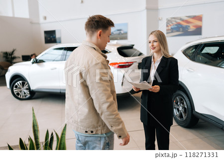 Attractive professional female car dealer in suit holding paper document, explaining buying new car to young man at modern showroom. Concept of choosing and buying new auto in dealership office. 118271331