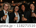 Group of women of different nationalities, dressed in business suits, sitting at bar and relaxing with beer after work. Cozy bar with soft warm light 118271510