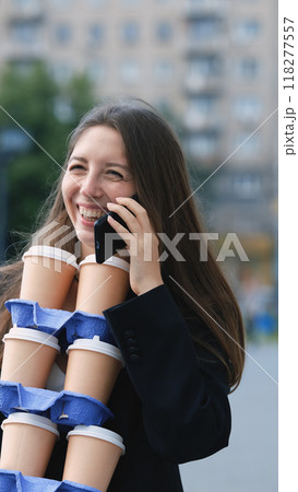A cute woman with long hair in an office black jacket carries many glasses of takeaway coffee in her hands and talks on her smartphone. 118277557