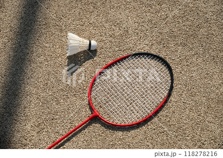 White badminton feather shuttlecock and badminton racket on beige floor sport badminton court in sunny shadow. Summer game outdoors. Close up. White badminton feather shuttlecock and badminton racket on beige floor sport badminton court in sunny shadow. Summer game outdoors. Close up. 118278216