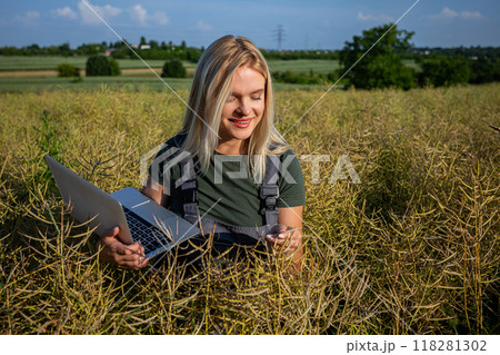 A smiling woman farmer checking the quality of rapeseed, standing at the rapeseed field and holding a laptop 118281302