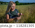 Female Farmer Gently Examining Mature Rapeseed Pods in a Field on a Clear Day with a Village in the Background 118281305