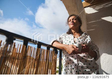 Woman enjoying morning coffee on sunny balcony in polka dot shirt, feeling relaxed and thoughtful Woman enjoying morning coffee on sunny balcony in polka dot shirt, feeling relaxed and thoughtful 118281675