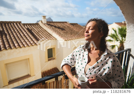 Woman enjoying a sunny morning on a balcony, feeling relaxed and peaceful while holding a cup of coffee, with charming Mediterranean architecture in the background. 118281676