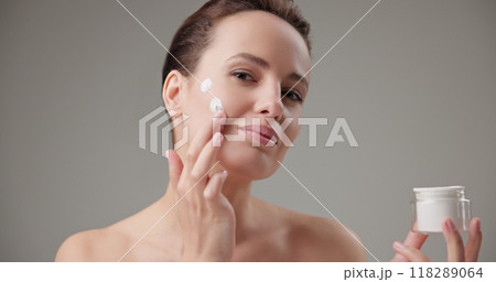 Beauty, health, cosmetics, anti-aging therapy and skin care concept - young beautiful brunette Caucasian woman holds a jar of moisturizer in her hands and applies the cream to her face skin Beauty, health, cosmetics, anti-aging therapy and skin care concept - young beautiful brunette Caucasian woman holds a jar of moisturizer in her hands and applies the cream to her face skin 118289064