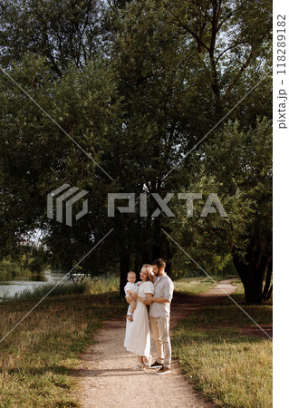 A family of three, a man and a woman with a child in their arms, stand in a park in the summer.  118289182