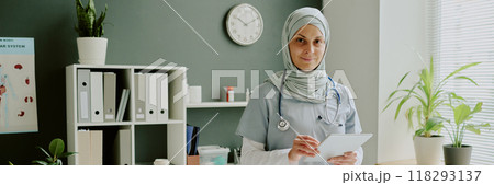 Portrait of smiling female doctor in modern office holding tablet and wearing professional attire including hijab standing by white shelves with medical files and folders Portrait of smiling female doctor in modern office holding tablet and wearing professional attire including hijab standing by white shelves with medical files and folders 118293137