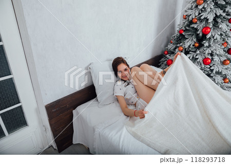 Woman in pajamas on bed in bedroom woke up by Christmas tree with gifts Woman in pajamas on bed in bedroom woke up by Christmas tree with gifts 118293718