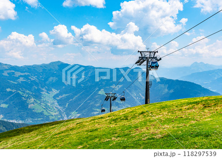 Visitors ride the cableway at Schmittenhohe, enjoying breathtaking views of the Austrian Alps on a sunny day. The lush green slopes contrast beautifully with the blue sky and distant mountains. 118297539