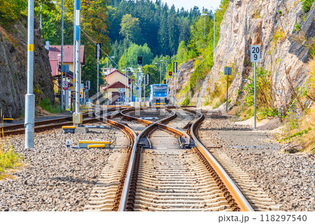 The railway tracks curve through Smrzovka station in Czechia, showing a train approaching in bright daylight. Surrounding greenery and buildings add to the picturesque setting. 118297540
