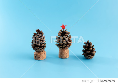 Three pine cones of different sizes on blue surface, mounted on cork bases, resembling tiny trees. Large pine cone with red star top, showcasing festive DIY craft with natural materials 118297979