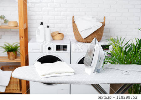 White Iron on Grey Ironing Board Next to Laundry Machines in White Room White Iron on Grey Ironing Board Next to Laundry Machines in White Room 118299552