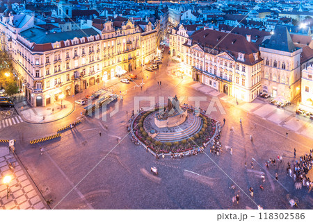 As evening falls, Old Town Square in Prague comes alive with vibrant lights, casting a warm glow on historic buildings and the bustling crowd gathered around the central fountain. 118302586