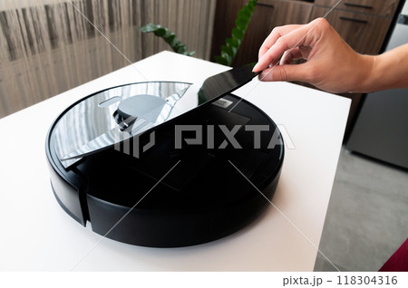 Modern robot vacuum cleaner after cleaning the apartment, a woman's hand opens the container lid to clean the vacuum cleaner after cleaning Modern robot vacuum cleaner after cleaning the apartment, a woman's hand opens the container lid to clean the vacuum cleaner after cleaning 118304316