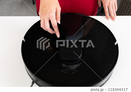Robot vacuum cleaner with round black body on white background table, new home assistant, woman hand reaches out to turn it on and check, close-up 118304317