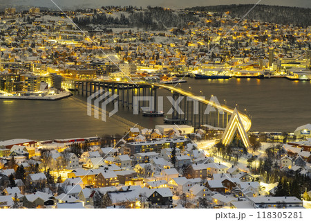 Panorama of Norwegian city of Tromso in the winter. Snowy roofs, bridge, embankment near the port and fishing ships. Evening lights 118305281