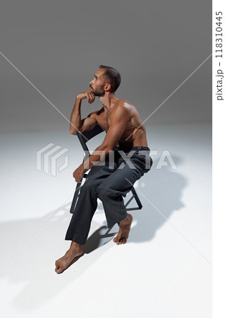 Thoughtful man, sitting on chair with one hand resting on his chin, strike contemplative pose while showcasing his toned, muscular physique. 118310445