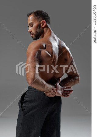 Rear view photo of young man with muscular body posing shirtless holding hands behind back against grey studio background. Rear view photo of young man with muscular body posing shirtless holding hands behind back against grey studio background. 118310455