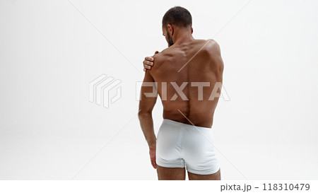 Athletic, shirtless man gently places his hand on his shoulder, emphasizing his muscular frame against white background. Athletic, shirtless man gently places his hand on his shoulder, emphasizing his muscular frame against white background. 118310479