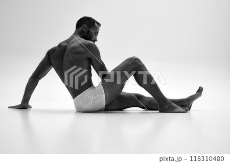 Young man sitting on floor, posing in underwear shows his muscular body against white background. Black and white photo. Young man sitting on floor, posing in underwear shows his muscular body against white background. Black and white photo. 118310480
